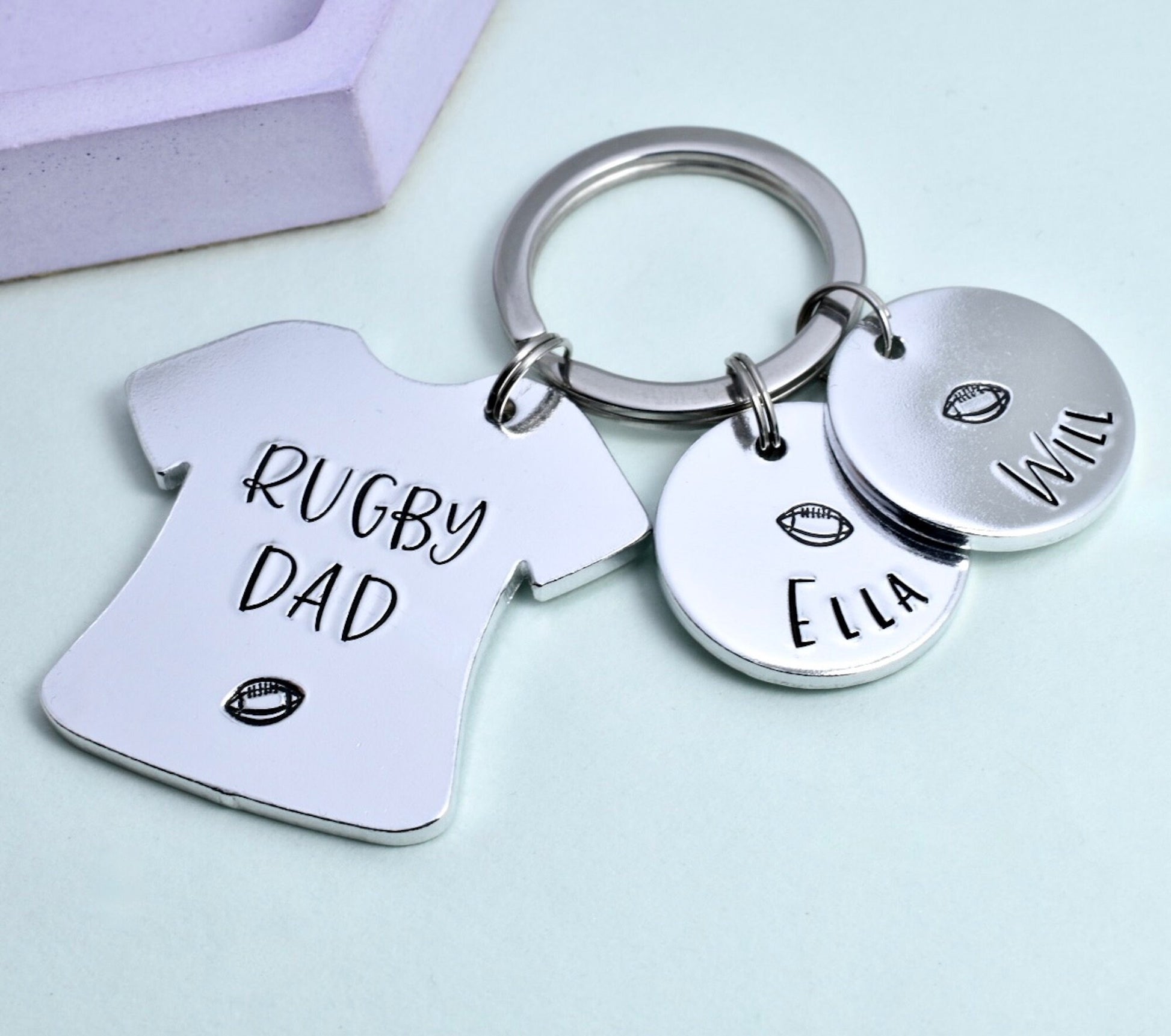 A personalised metal keyring with a silver shirt design that reads 'RUGBY DAD' and two smaller discs with names 'Ella' and 'Will'. The keyring is split into three parts, connected by a split ring.