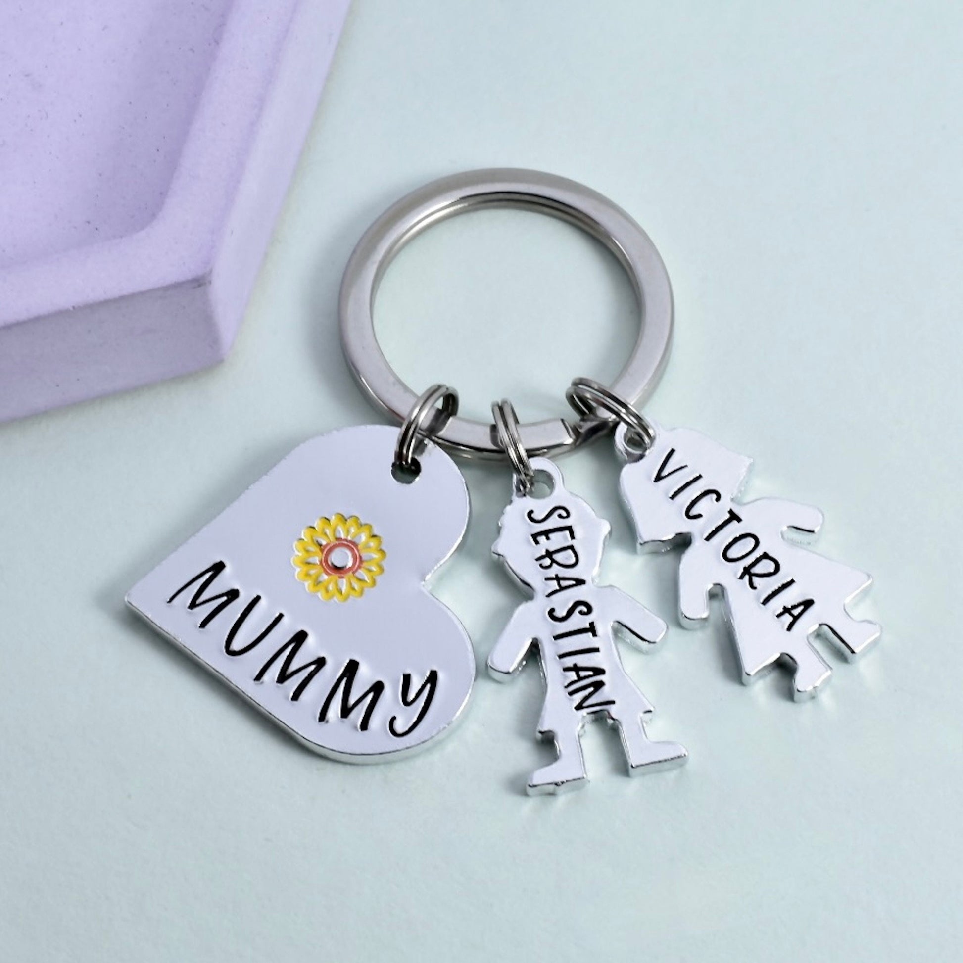 A personalised aluminum keyring with a heart-shaped charm that has a yellow flower design, and two other charms shaped as people. The keyring is inscribed with the word 'MUMMY' and the names 'Victoria' and 'Sebastian'.