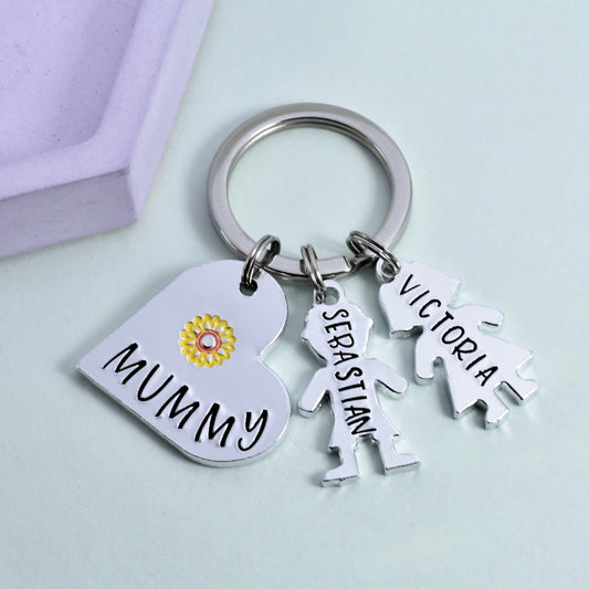 A personalised aluminum keyring with a heart-shaped charm that has a yellow flower design, and two other charms shaped as people. The keyring is inscribed with the word 'MUMMY' and the names 'Victoria' and 'Sebastian'.
