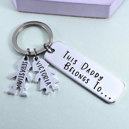 A personalised keyring with a rectangular tag and two metallic charms shaped as individuals, attached to a keyring. The rectangular tag has the inscription 'This Daddy Belongs To.