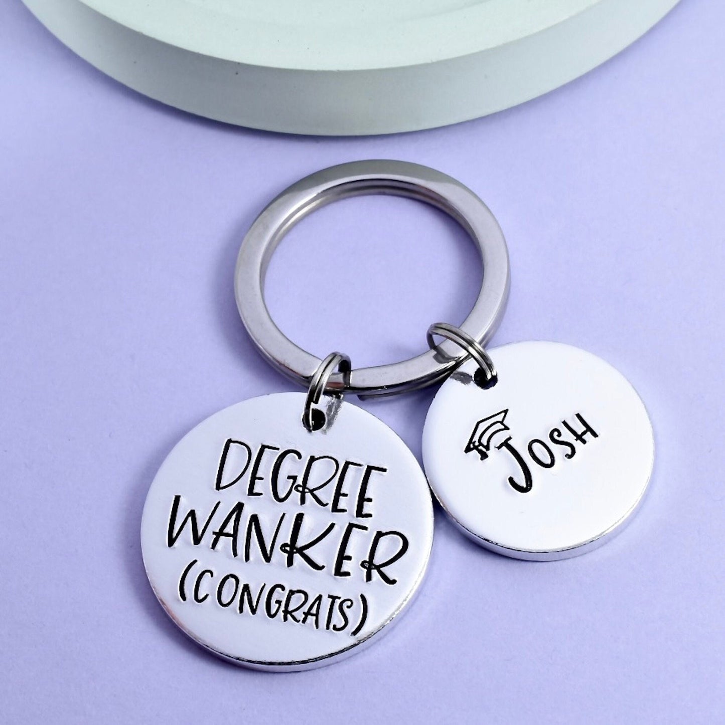 A personalised graduation keyring with the text 'DEGREE WANKER' on a smaller circular disc, and the name 'Josh' on a smaller disc attached to it, all on a light background.