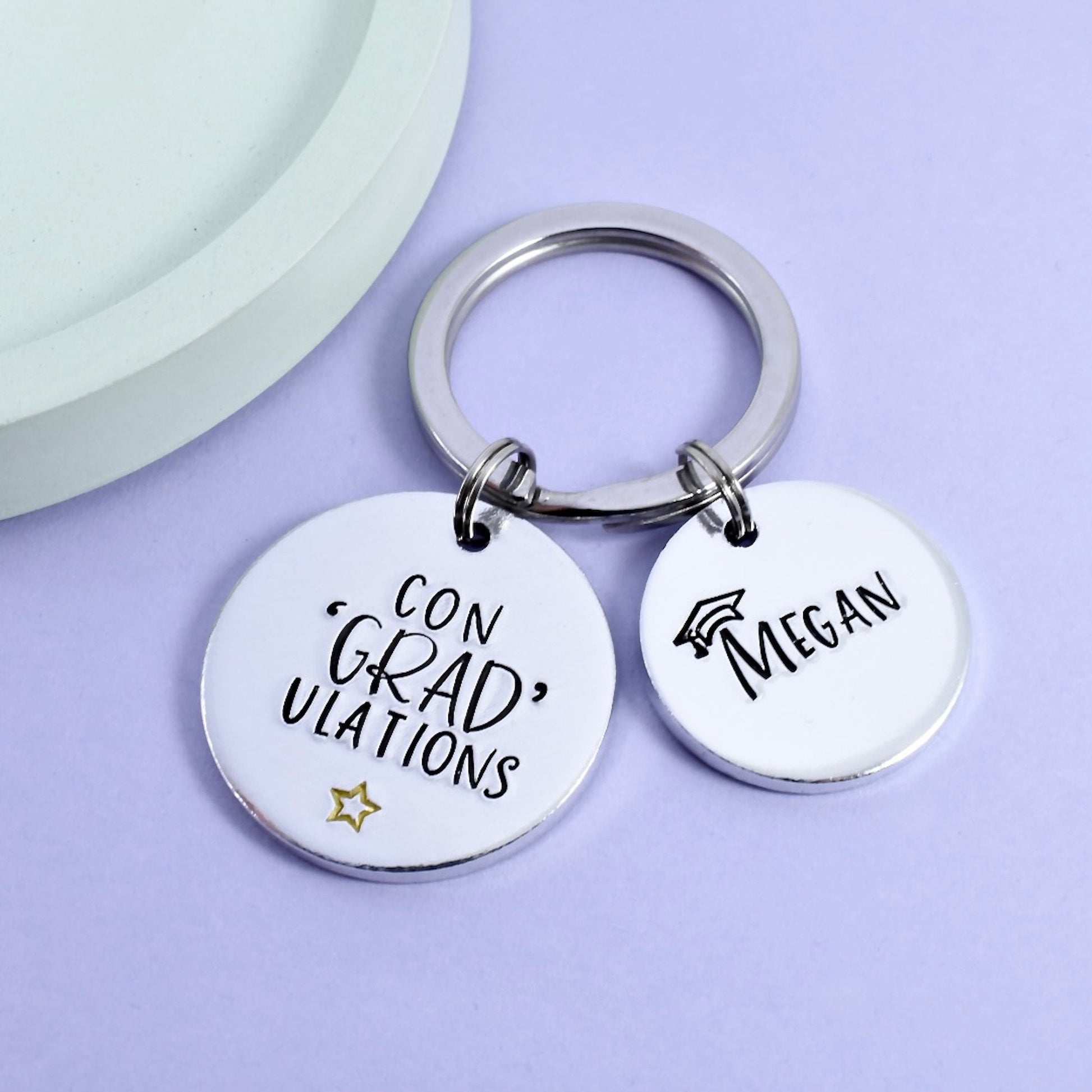 A personalised graduation keyring with two round metal discs, one with the text 'CON GRAD ULATIONS' and the other with a name, attached to a keyring. The keyring and the discs are silver-colored.