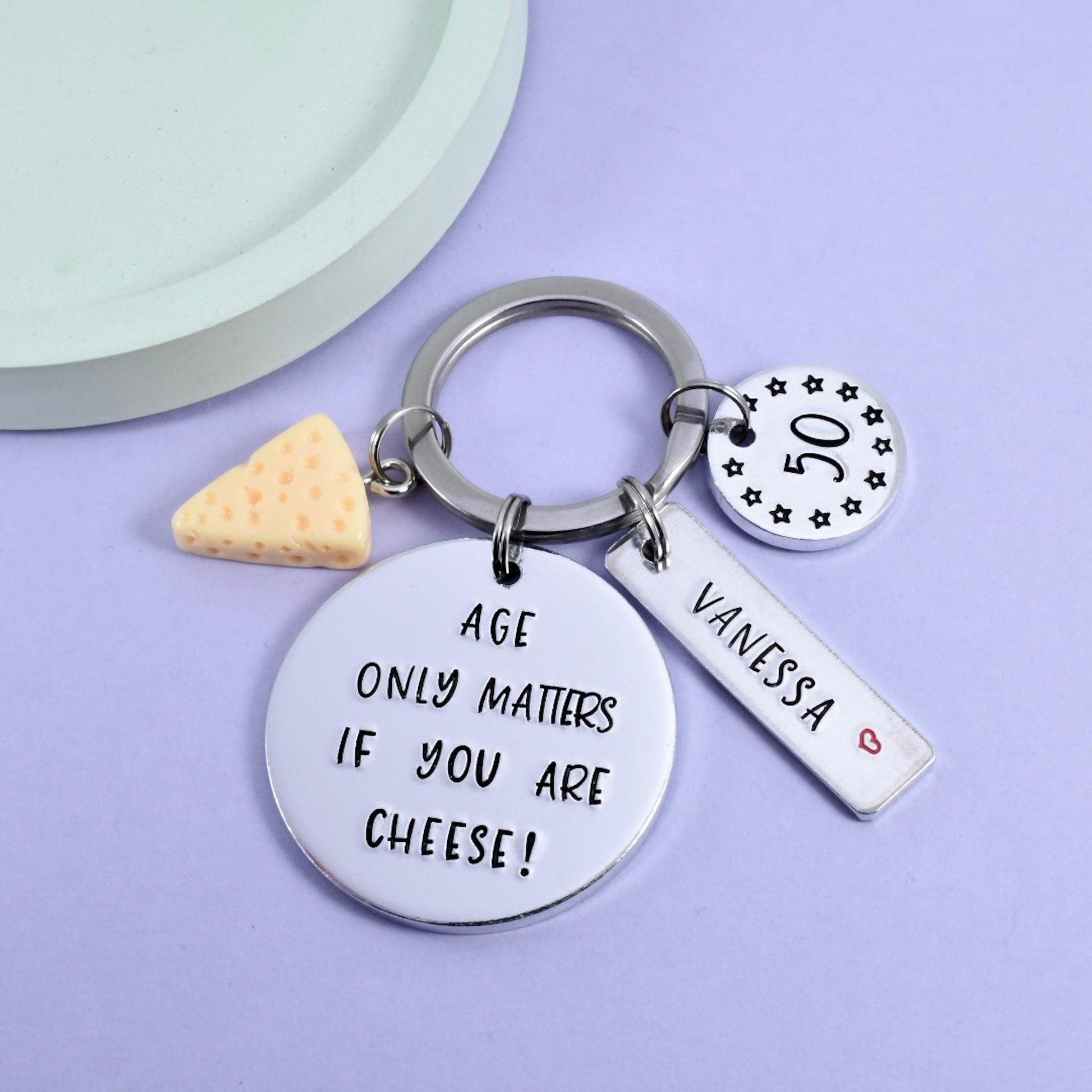 A personalised keyring with round and rectangular metal discs, one shaped like a cheese wedge, and a split ring. The discs are stamped with "age only matters if you are cheese!" and age and the rectangular tag has a name.