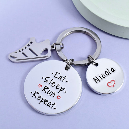 A personalised keyring with a circular disc, split ring, and trainer charm. The disc has text 'EAT, SLEEP, RUN, REPEAT' and a runner's name, NICOLA, with a heart symbol between each word.