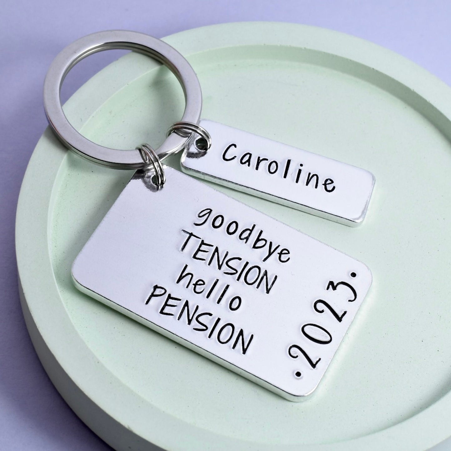 A personalised retirement keyring with two aluminium tags. One tag has the text 'goodbye TENSION hello PENSION' along with the year, and the other tag is personalised with a name.