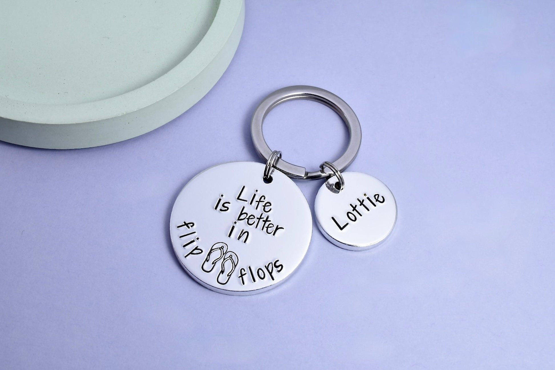 A personalised keyring with a split ring, featuring a large disc with the inscription 'LIFE IS BETTER IN FLIP FLOPS' and the recipient's name 'Lottie' on it, placed on a light background.