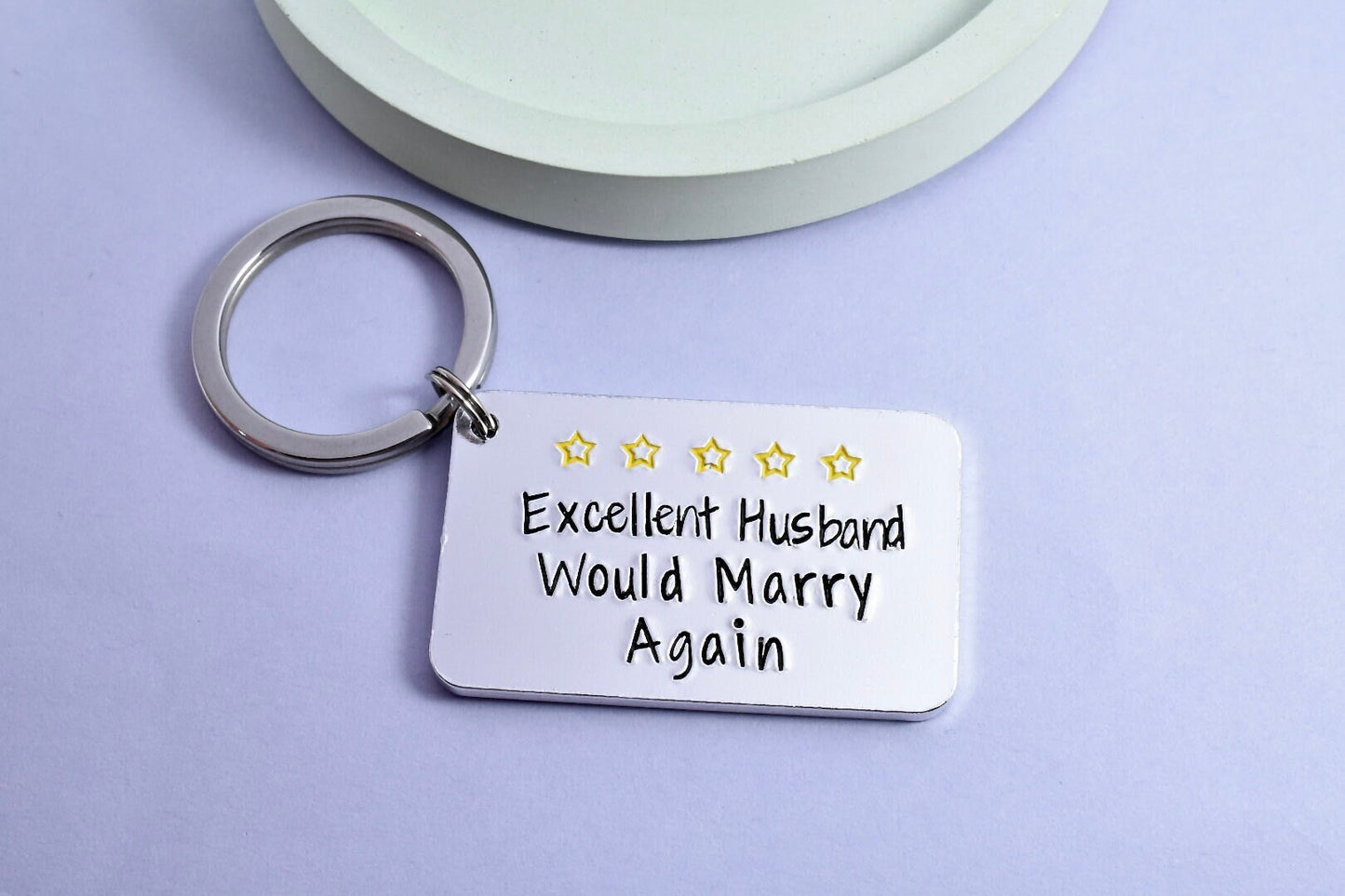 A silver-colored keyring with the inscription 'Excellent Husband, Would Marry Again' and five gold stars, placed on a light background.
