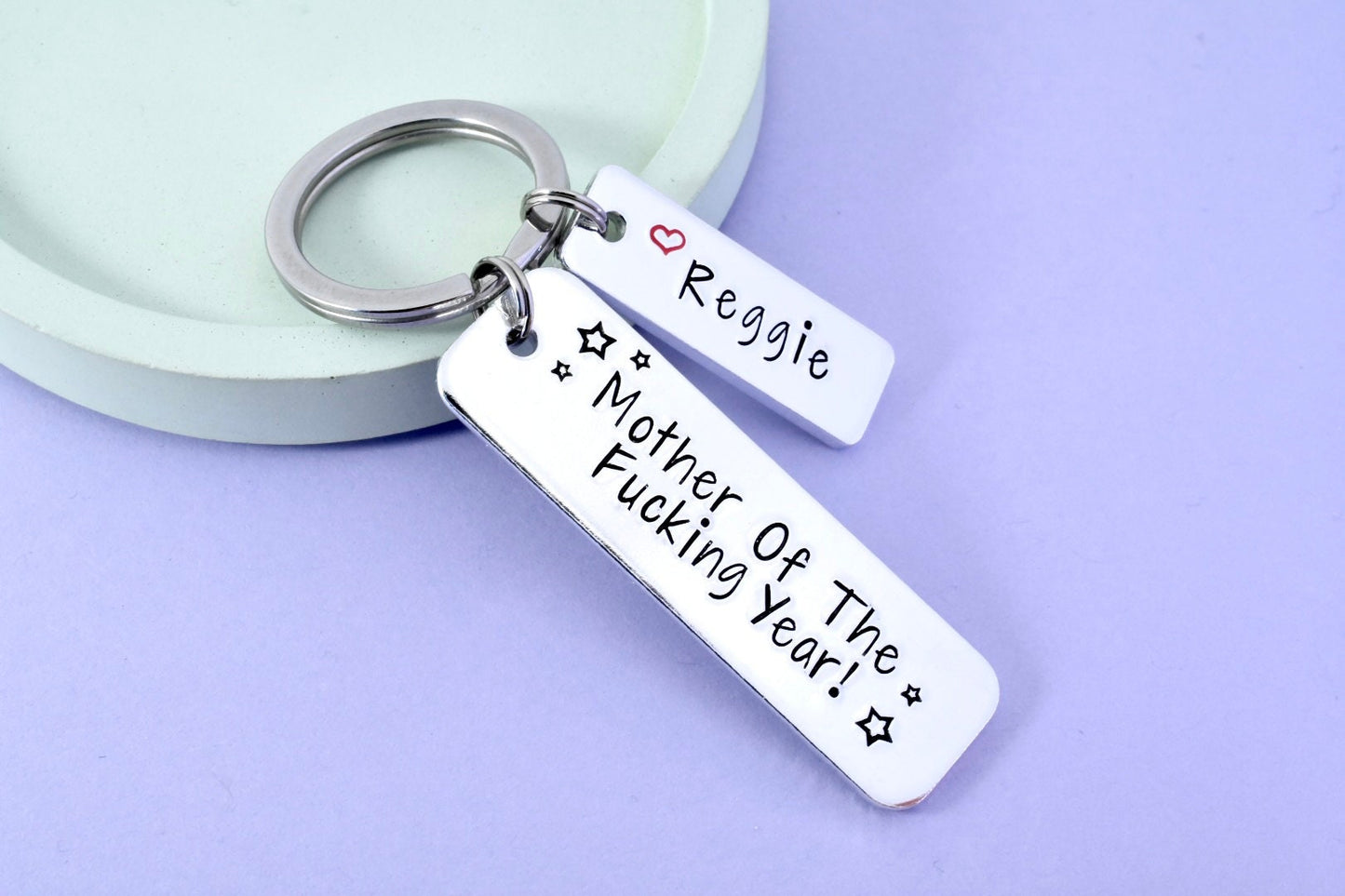A silver-colored keyring with the inscription 'MOTHER OF THE FUCKING YEAR!' and a small heart symbol, displayed on a light background.
