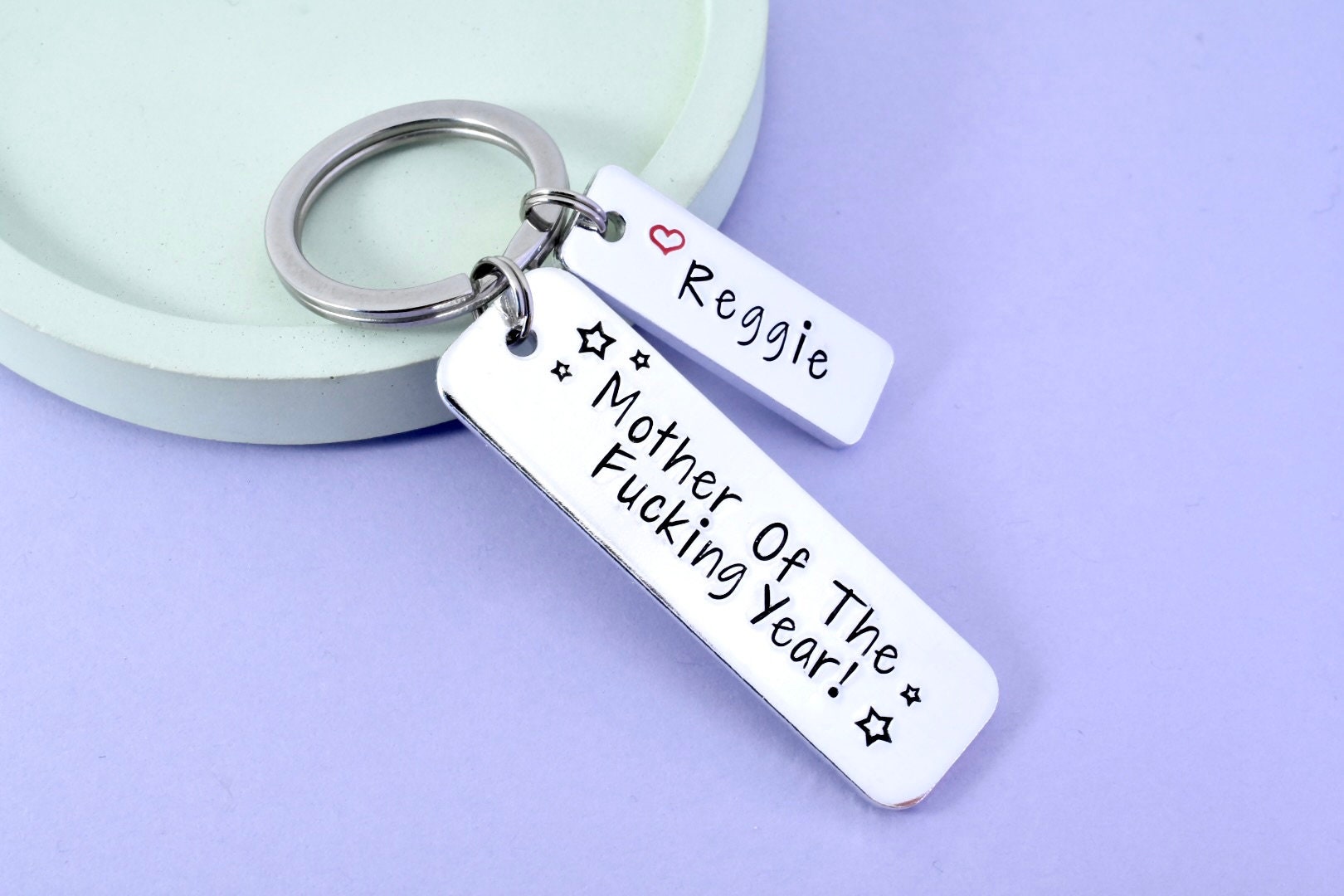 A silver-colored keyring with the inscription 'MOTHER OF THE FUCKING YEAR!' and a small heart symbol, displayed on a light background.