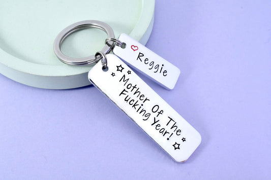 A silver-colored keyring with the inscription 'MOTHER OF THE FUCKING YEAR!' and a small heart symbol, displayed on a light background.