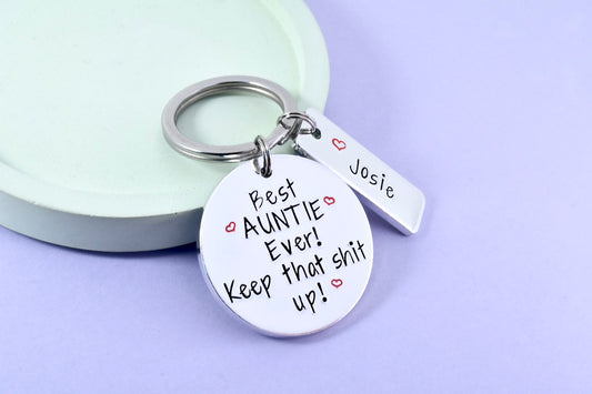 A personalised keyring with a large aluminum disc that says 'Best AUNTIE Ever! Keep that shit up!' and a smaller rectangle that has names. 