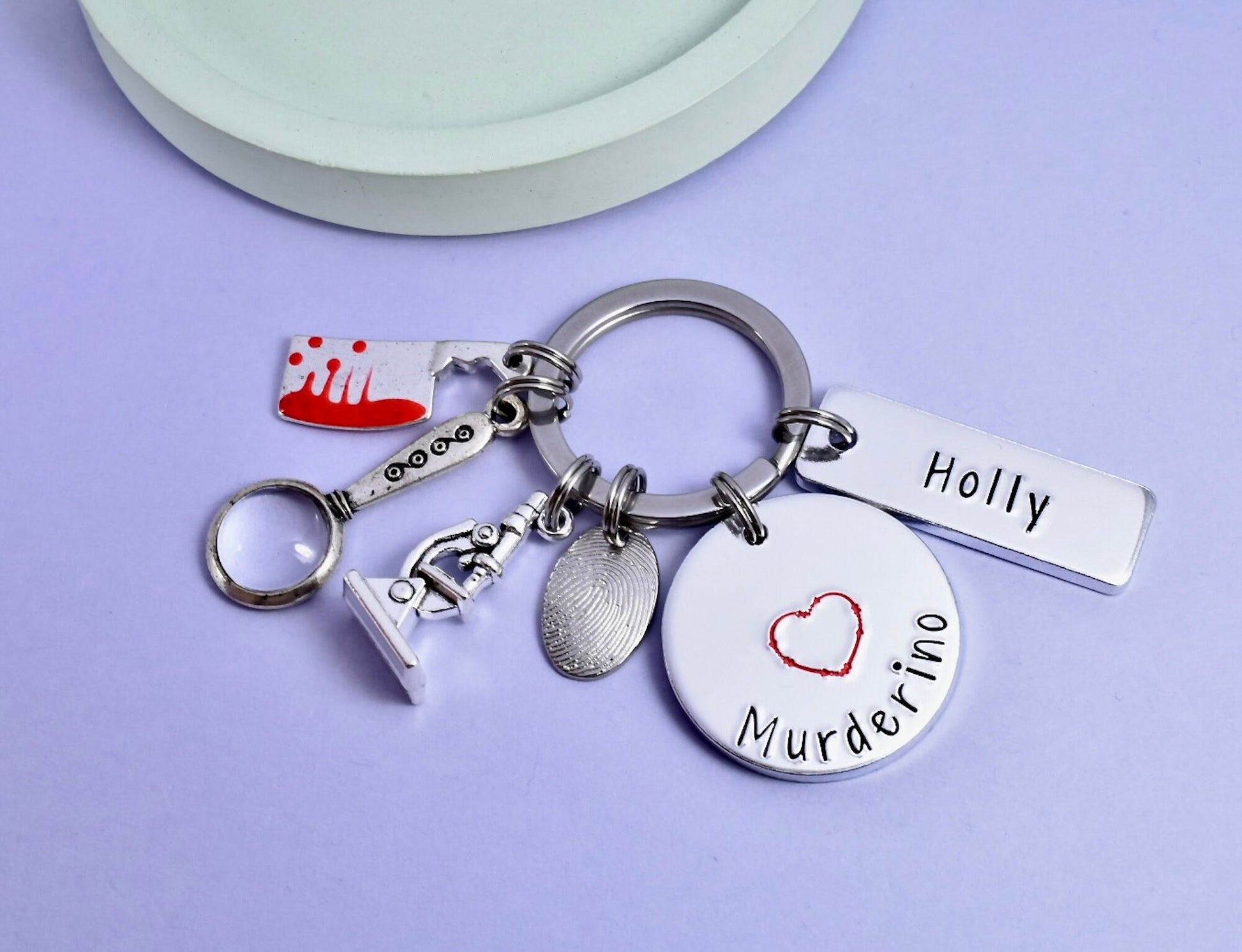A personalised keyring with a 'Murderino' label, true crime inspired charms including a fingerprint, microscope, magnifying glass, and a bloodied meat cleaver, and a custom name 'Holly'.
