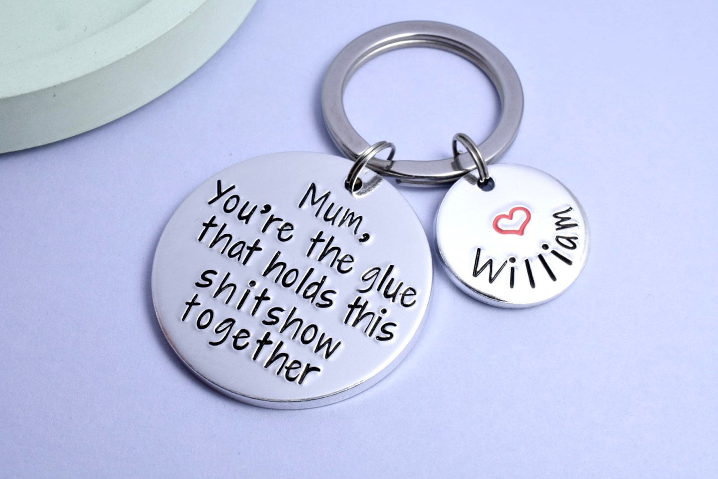 A silver-colored keyring with two discs, one larger with the text 'YOU'RE THE GLUE THAT HOLDS THIS SHITSHOW TOGETHER' and a smaller one attached customised with a red heart and name