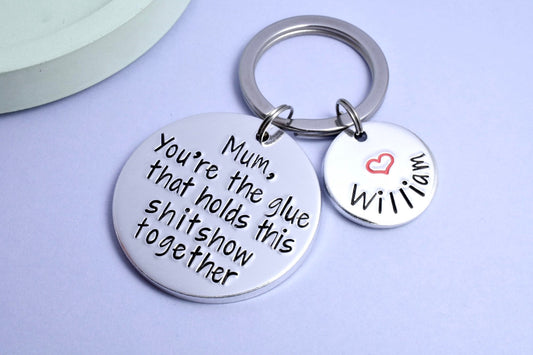 A silver-colored keyring with two discs, one larger with the text 'YOU'RE THE GLUE THAT HOLDS THIS SHITSHOW TOGETHER' and a smaller one attached customised with a red heart and name