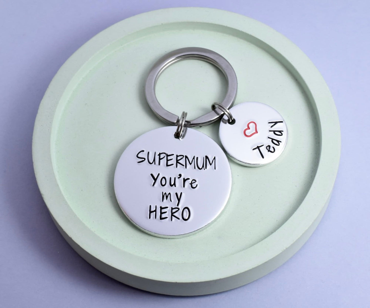 A personalised keyring with an aluminium disc stamped with 'SUPERMUM You're My HERO' alongside a smaller disc with a heart and a name. 

